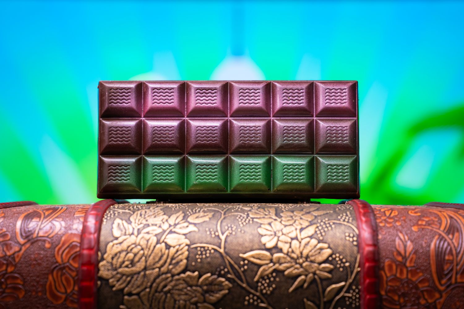 Rectangular chocolate bar decorated with shimmering shades of pink, purple, and green in front of an abstract blue and green backdrop. The chocolate is perched on top of an antique book. 